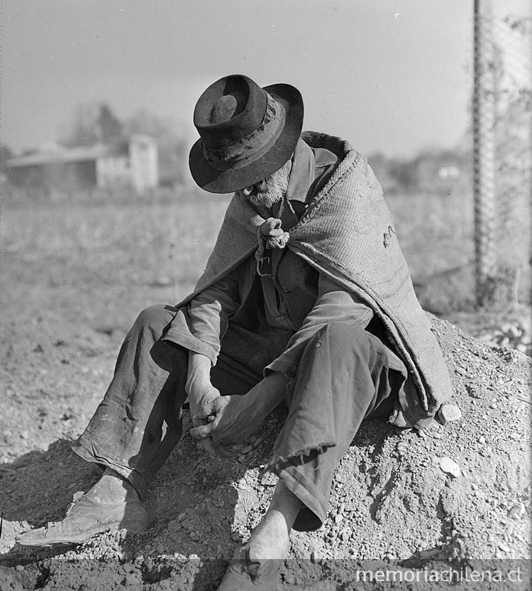 Hombre anciano de manos cruzadas y cabizbajo, con sombrero, sentado sobre un cerro de tierra