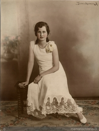 Adolescente de vestido blanco largo adornado con broderí en la parte inferior sentada en un piso de madera sobre alfombra, entre 1930 y 1940
