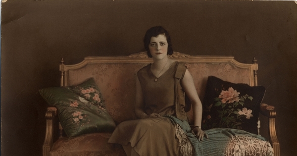 Mujer joven sentada en un sofá, con vestido café y mantilla sobre el regazo, entre 1930 y 1940