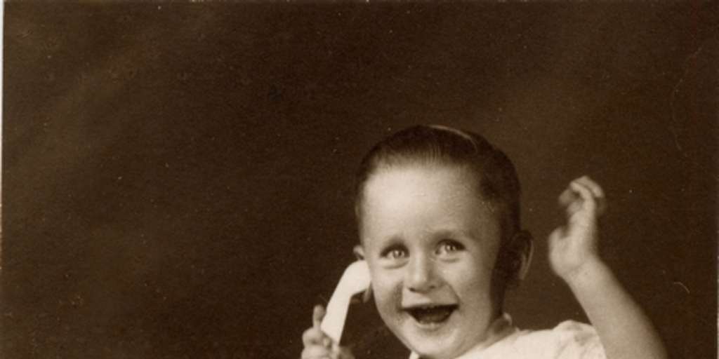 Niño sonriente sentado sobre un felpudo con pantalones cortos, camisa blanca manga corta, corbatín a rayas y chalas blancas, simulando hablar por un teléfono de juguete, 1950